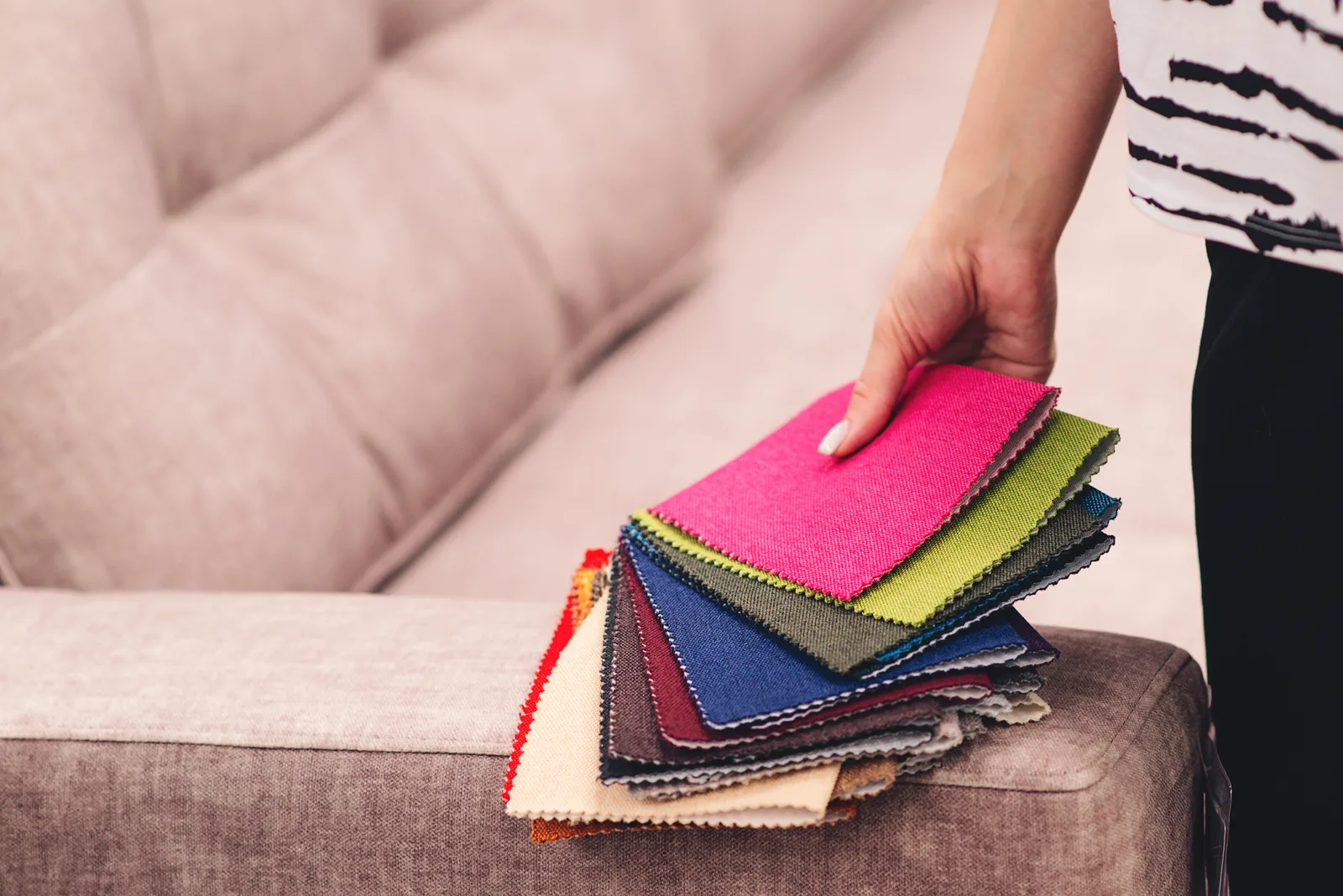 Hand holding a fan of upholstery fabric swatches over the arm of a sofa, the kind of fabric identification R&L Carpet Cleaning does before cleaning furniture in Kansas City homes