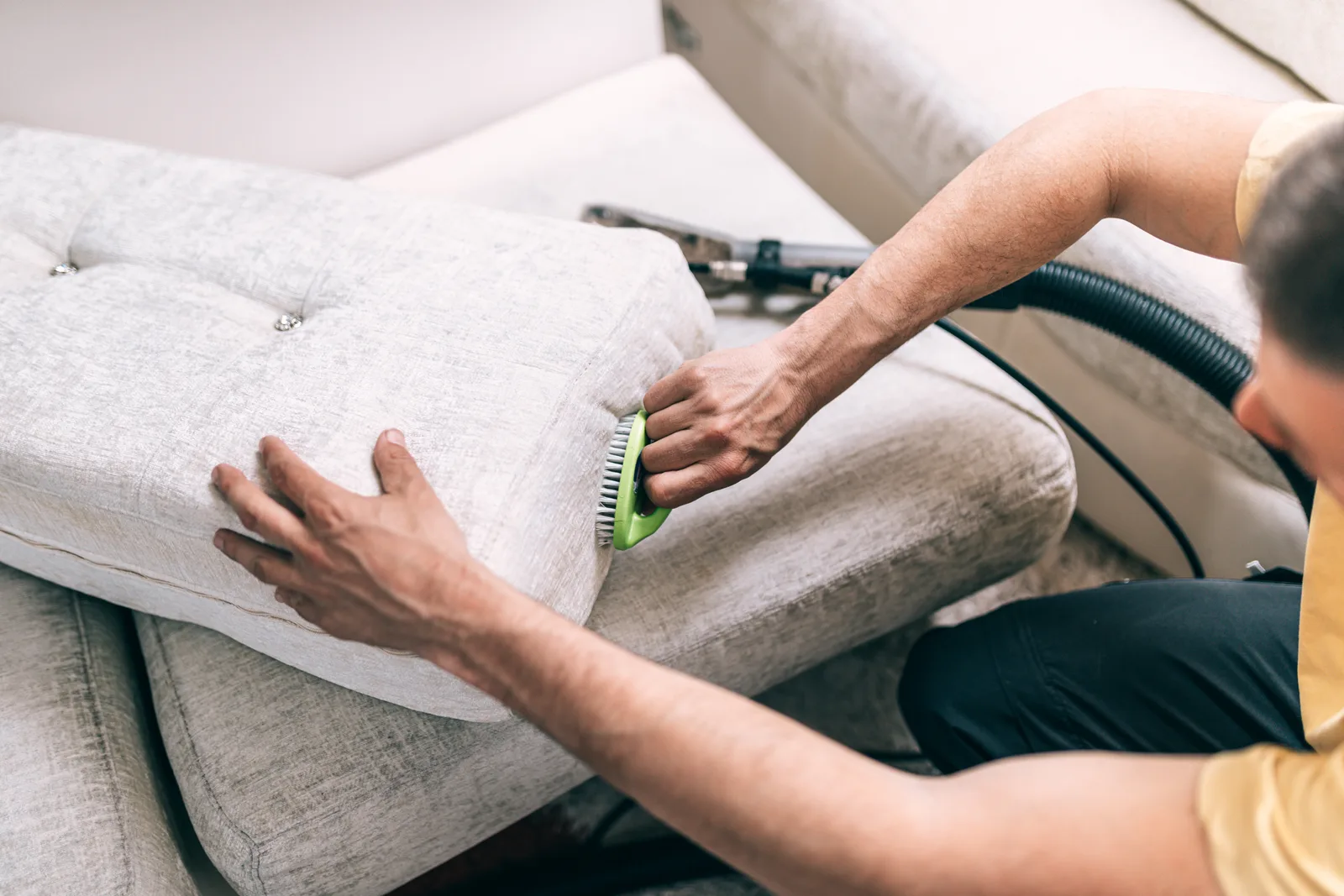 Upholstery cleaning technician using a hand extraction tool on a light gray fabric sofa cushion in a living room